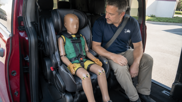 NEMT driver seated beside a pediatric mannequin secured in a child restraint system inside a red Dodge Grand Caravan